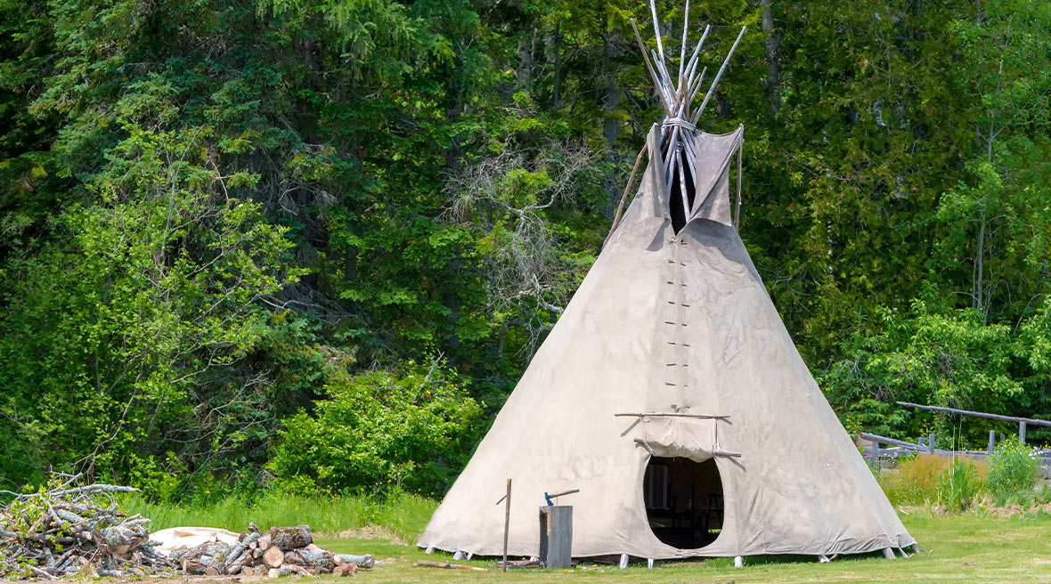 Tepee tent in a field with the door open surrounded by trees with an unlit fire to the left of it